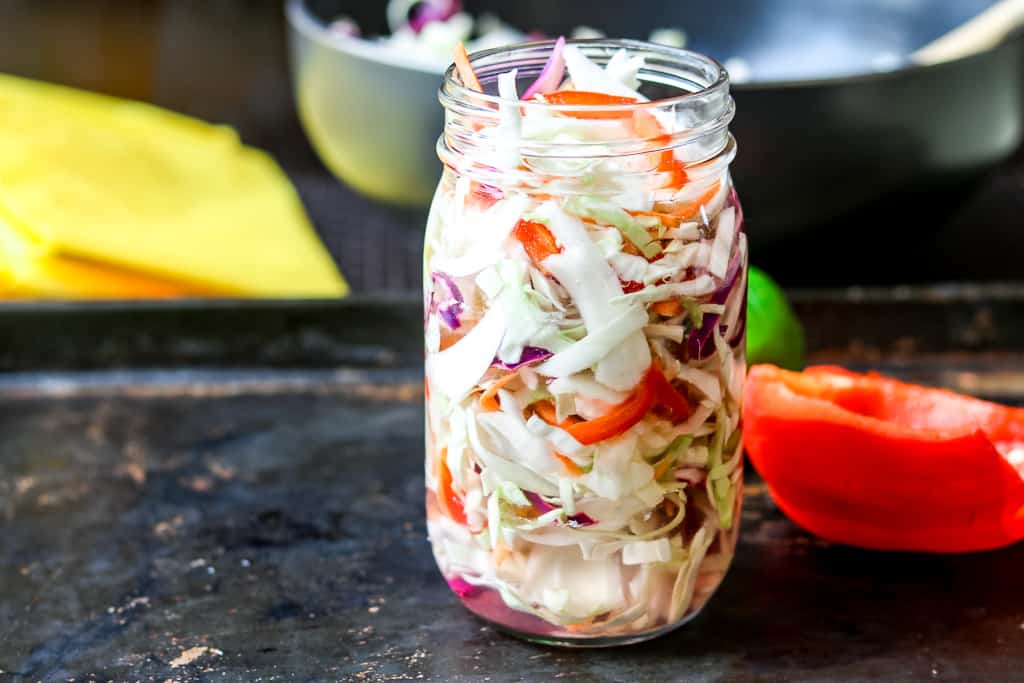 Haitian pikliz Recipe in a clear mason jar with red bell pepper in the back, yellow pepper and black bowl in the background.