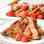 Two slices of French brioche French toast. Topped with Maple syrup and raspberries on a white plate with slices brioche French toast in the background.
