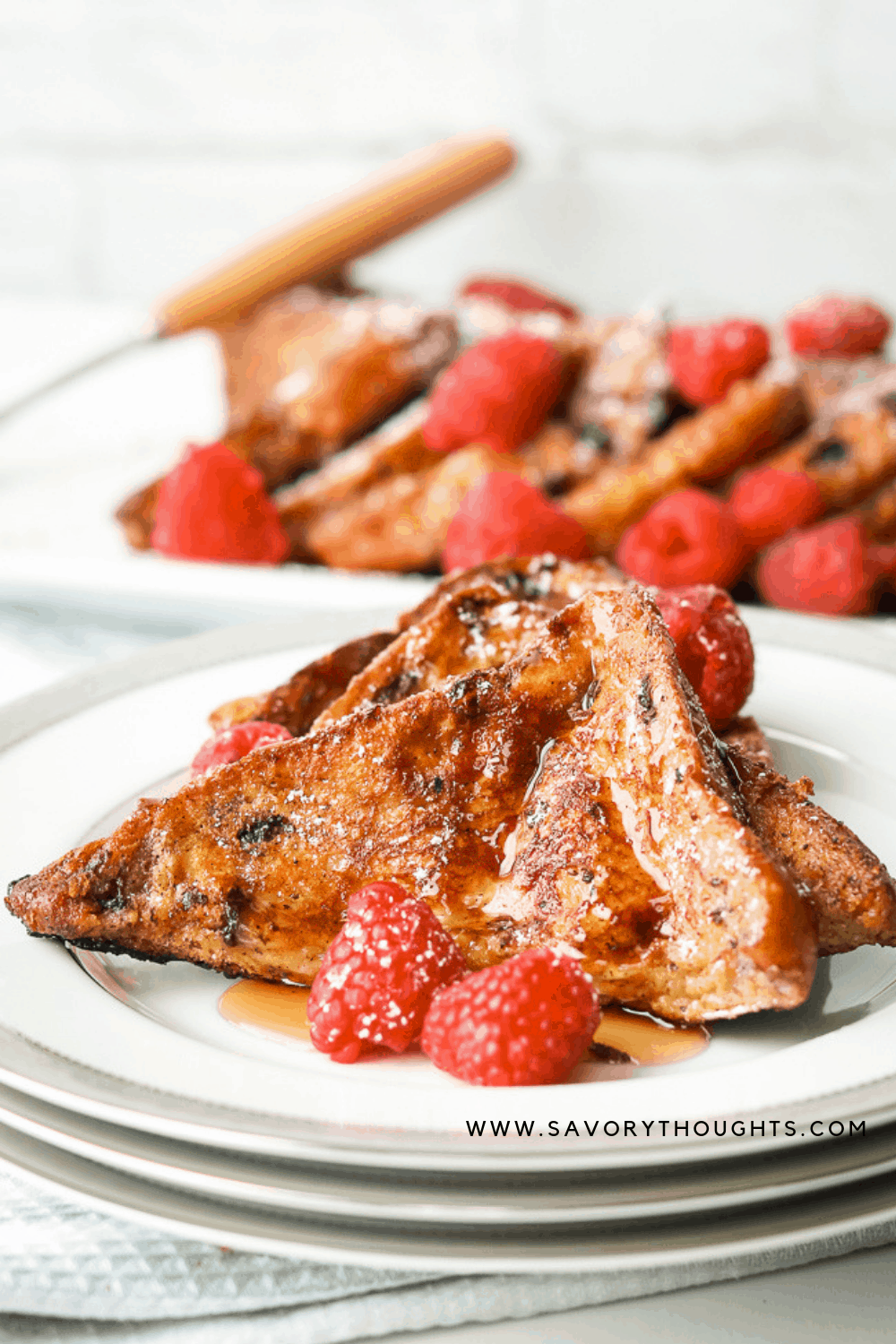 Two slices of French brioche French toast. Topped with Maple syrup and raspberries on a white plate with slices brioche French toast in the background.