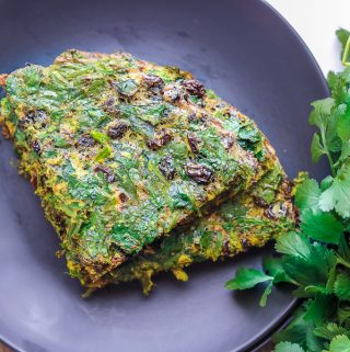 two pieces of Kuku Sabzi – Persian Herb Frittata on a black plate with fresh herbs on the side.