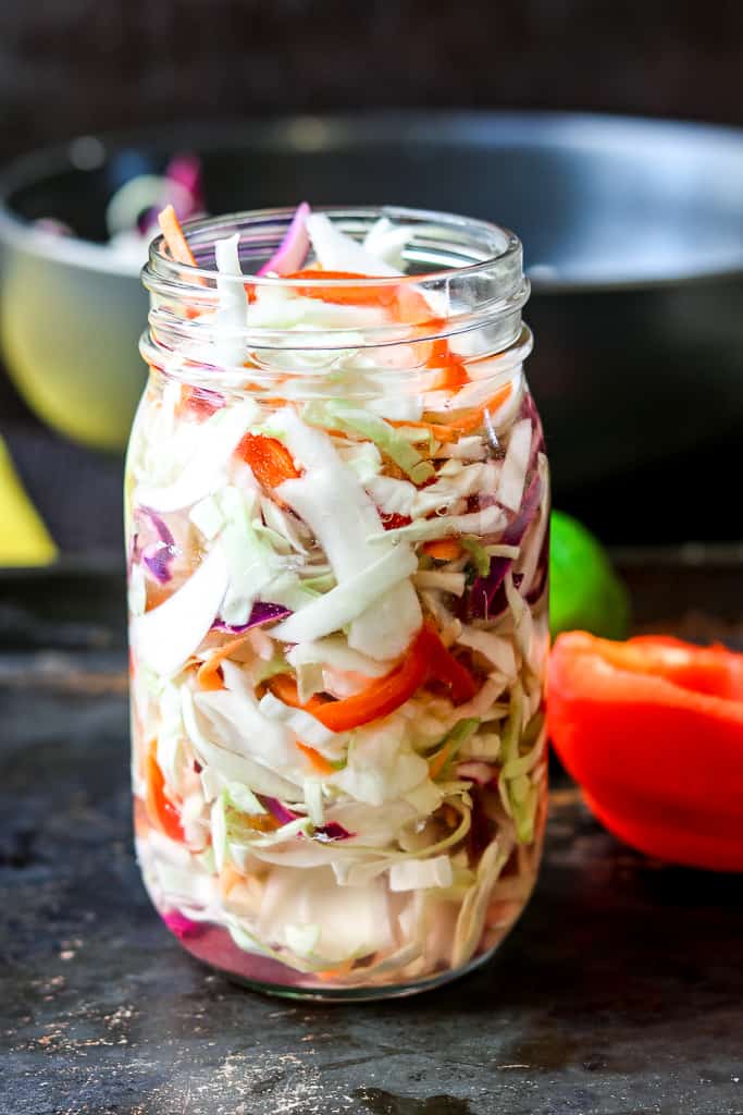 Haitian Pikliz Recipe in a mason jar with red bell pepper in the back, black bowl, and lime.