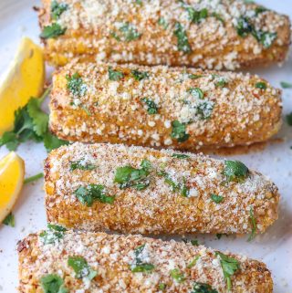 Four Mexican Street Corn (Elotes) in a roll on a white plate with two lemon wedges in the back. Topped with cheese and cilantro.