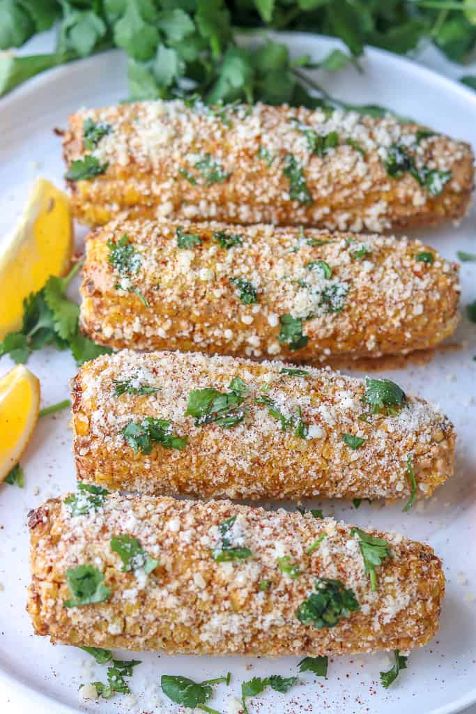 Four Mexican Street Corn (Elotes) in a roll on a white plate with two lemon wedges in the back. Topped with cheese and cilantro.