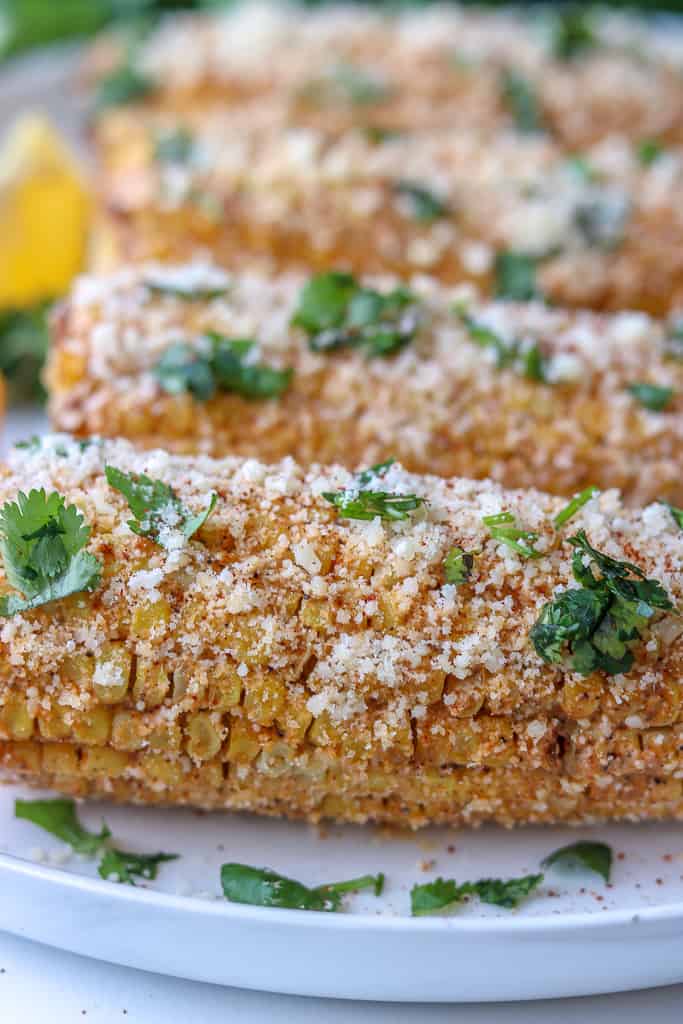 Closeup picture of the Mexican corn on the cob topped with cotija cheese and chopped cilantro. the picture shows 4 corn on the cob on a white plate. Sprinkled with chopped cilantro.