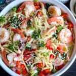 scampi pasta recipe on a white plate, topped with shrimp, herbs, tomatoes, and onions.