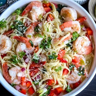 scampi pasta recipe on a white plate, topped with shrimp, herbs, tomatoes, and onions.