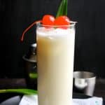 Pina-Colada-Mocktail-Coco-Colada in a tall high glass with two cherries on top and cocktail mixture in the background.