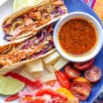 Haitian chicken tacos on a blue and white plate, topped with pikliz accompanied with marinated tomatoes, Swiss cheese, lime wedge, taco and Haitian sauce, pikliz.