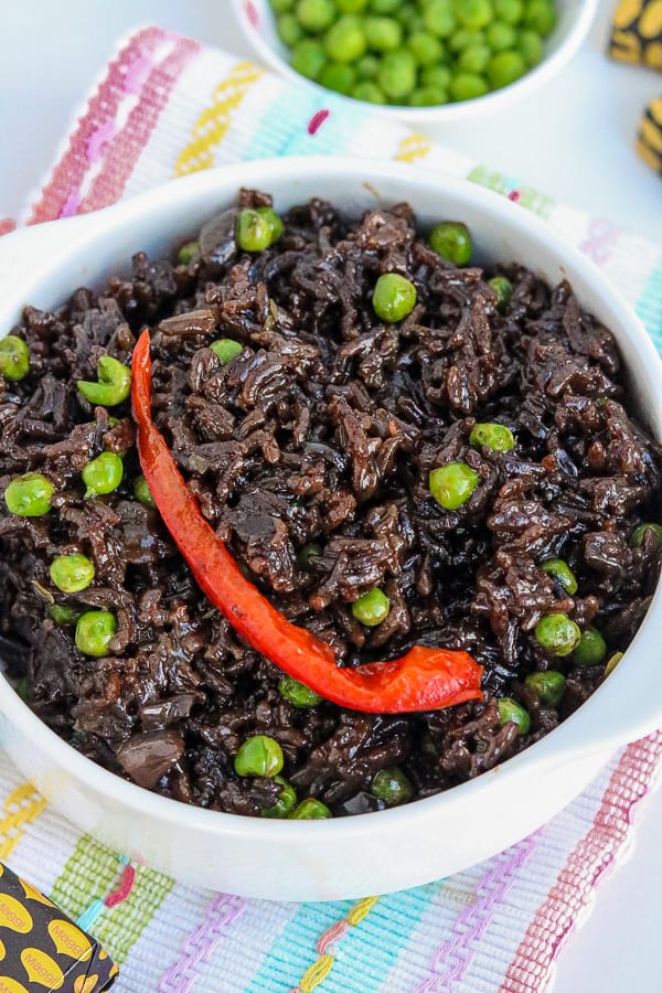 Haitian Black Rice in white bowl topped with red bell pepper and green peas.