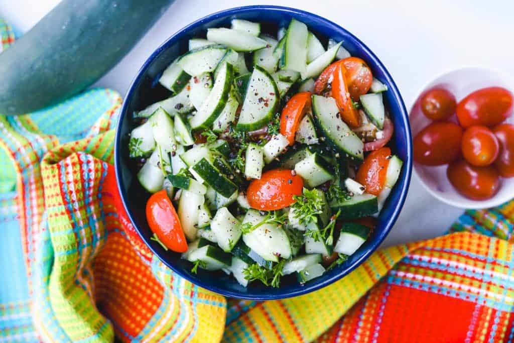 Cucumber Salad in blue bowl with cherry tomatoes on the side and colorful dishcloth