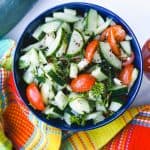 Cucumber Salad in blue bowl with cherry tomatoes on the side and colorful dishcloth