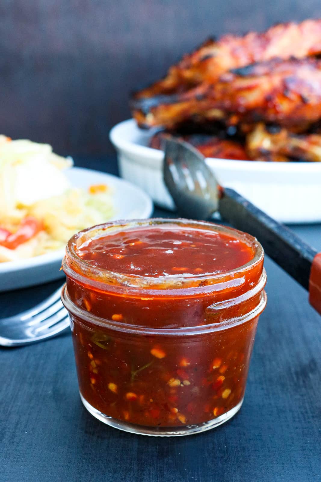 BBQ Sauce in a glass jar on black table with meat and vegetables in white plate in the background