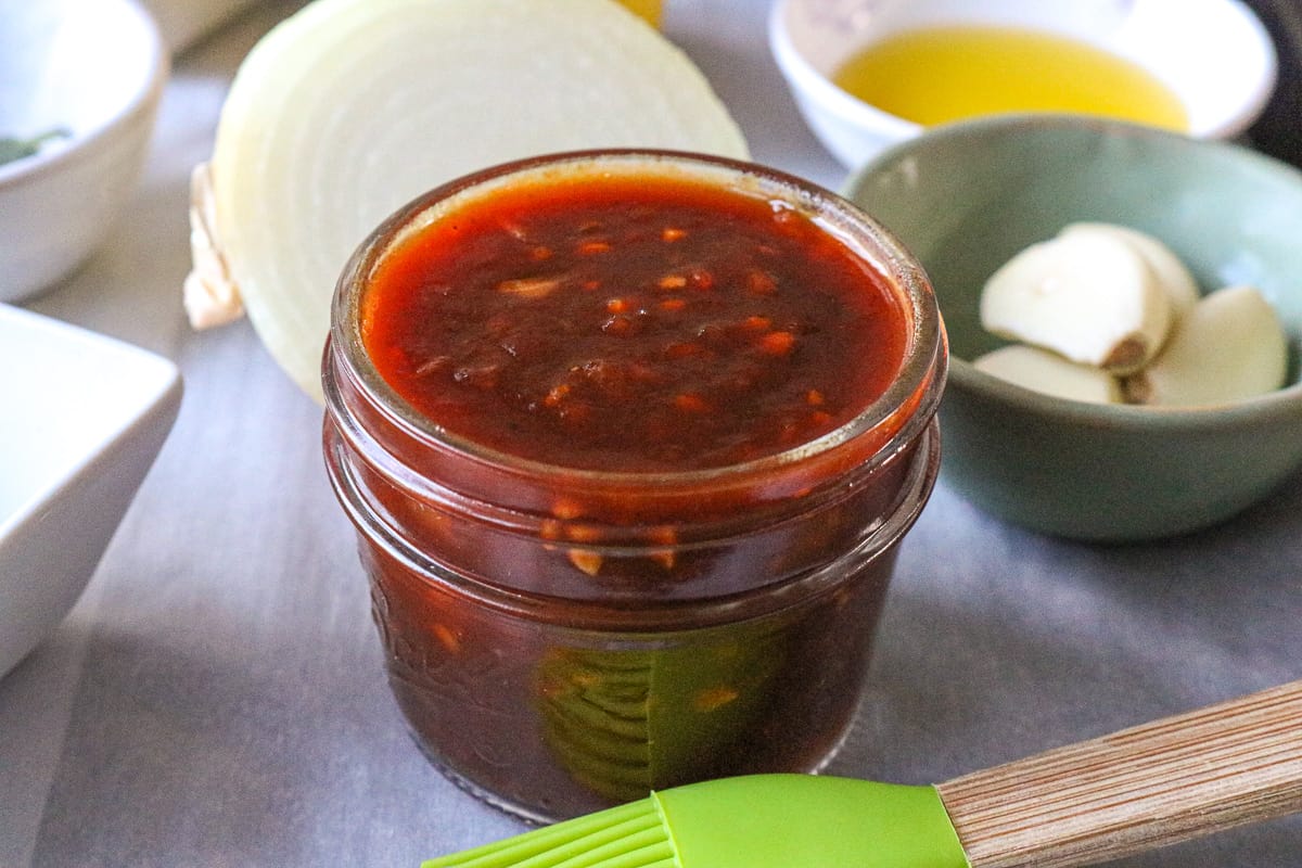 Sauce in glass jar with garlic, onion, and oil in the background.