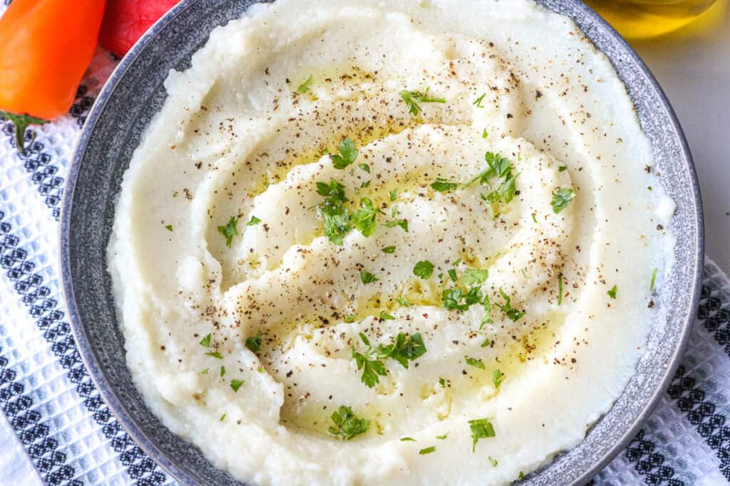 cauliflower in mashed formed in gray bowl. topped with herbs and olive oil.