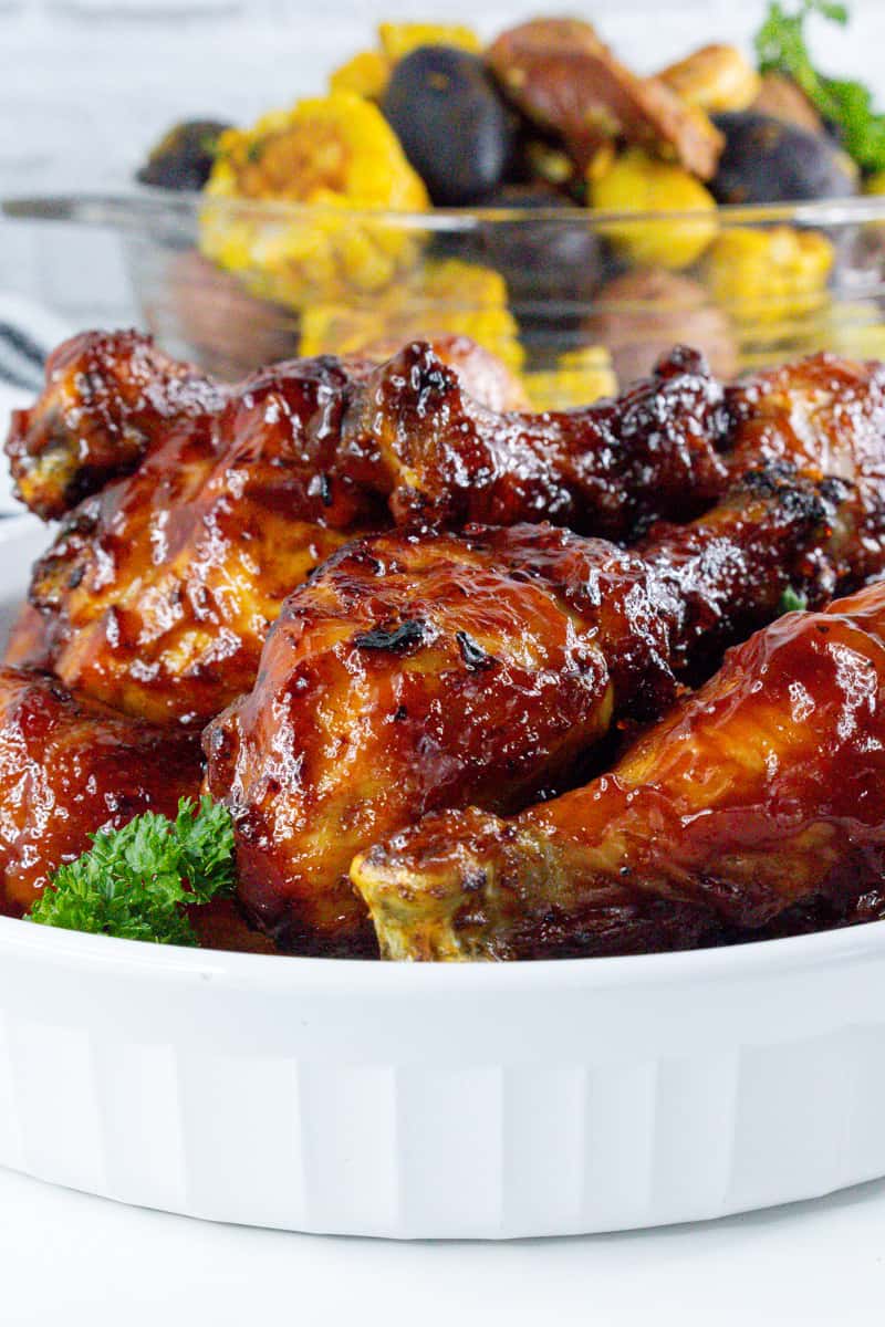 cooked bbq air fryer drumsticks in white bowl with parsley and corn, potatoes in the back