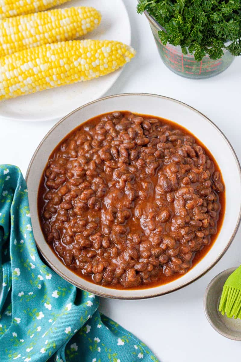 vertical view of baked beans in brown bowl with corn in the background and parsley