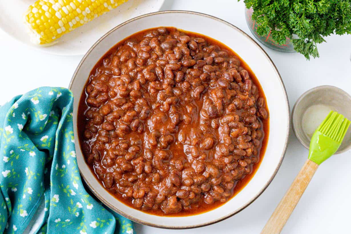 baked beans in brown bowl with corn in the background and parsley