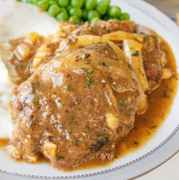 Salisbury steak recipe topped with onions in and gravy in white plate with mashed potatoes and green peas