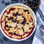 fully baked strawbetry blueberry pie in white pie dish with blueberries and a kitchen towel on the side