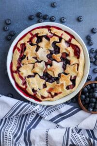 fully baked strawbetry blueberry pie in white pie dish with blueberries and a kitchen towel on the side