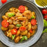 homemade Pesto gnocchi topped with tomato, pine nuts, and fresh basil in gray bowl.