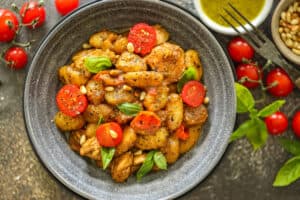homemade Pesto gnocchi topped with tomato, pine nuts, and fresh basil in gray bowl.
