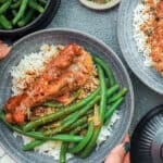 Hands holding Smothered turkey wings in baking dish topped with red pepper flakes and dried parsley over a bed of rice and green beans