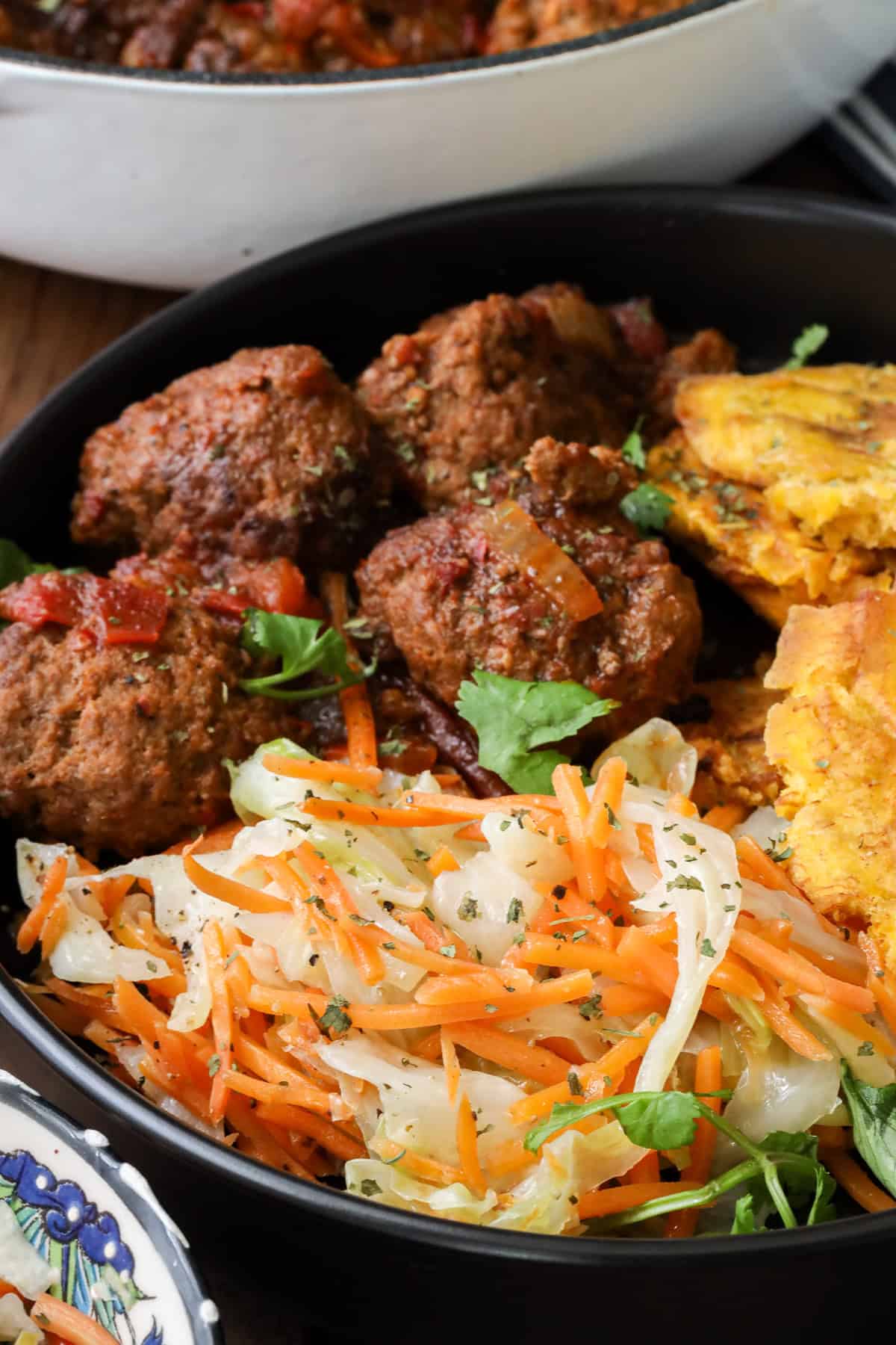 Steamed cabbage Jamaican Style prepared with carrots and Harissa meatballs and Haitian Fried Plantains in black plate