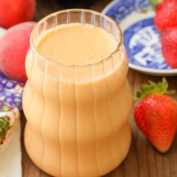 Strawberry Peach Smoothie in clear glass with strawberries and peach on a wooden board