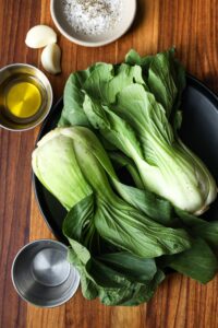2 baby Bok Choy, olive oil, water, and salt and paper on a wooden board.