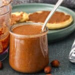 Smooth roasted homemade almond butter in a glass jar with almonds scattered around