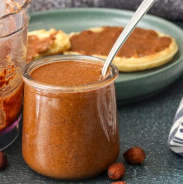 Smooth roasted homemade almond butter in a glass jar with almonds scattered around